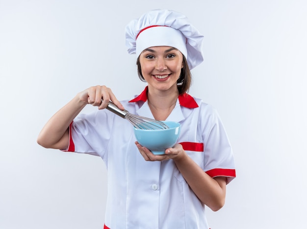 Sonriente sonriente joven cocinera vistiendo uniforme de chef sosteniendo el tazón con batidor aislado sobre fondo blanco.