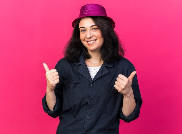 Sonriente sonriente joven mujer caucásica del partido vistiendo gorro de fiesta mirando al frente mostrando los pulgares para arriba aislado en la pared rosa