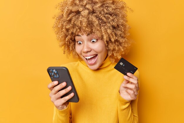 Sonriente, sorprendida, joven de cabello rizado mira fijamente el teléfono inteligente que sostiene la tarjeta de crédito, hace compras en línea, proporciona pagos por Internet, transfiere dinero en la aplicación ebank, posa en el interior contra la pared amarilla