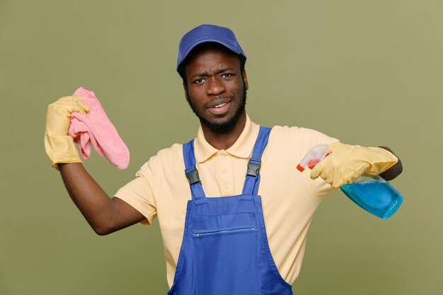 sonriente sosteniendo agente de limpieza con trapo joven limpiador afroamericano macho en uniforme con guantes aislado sobre fondo verde