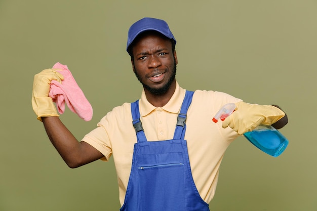 sonriente sosteniendo agente de limpieza con trapo joven limpiador afroamericano macho en uniforme con guantes aislado sobre fondo verde