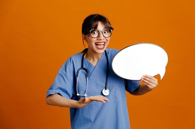 Sonriente sosteniendo la burbuja del habla joven doctora vistiendo uniforme fith estetoscopio aislado sobre fondo naranja