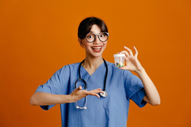 Sonriente sosteniendo un contenedor de pastillas, una joven doctora con un estetoscopio uniforme aislado en un fondo naranja