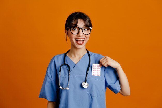 Sonriente sosteniendo píldoras joven doctora vistiendo uniforme fith estetoscopio aislado sobre fondo naranja