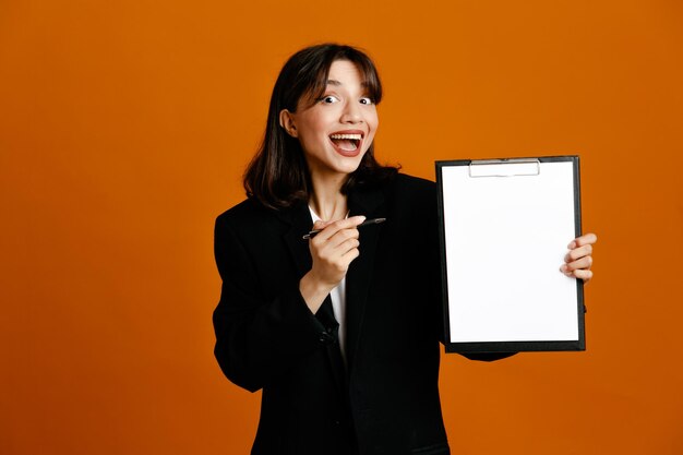 Sonriente sosteniendo portapapeles y pluma joven hermosa mujer vistiendo chaqueta negra aislado sobre fondo naranja