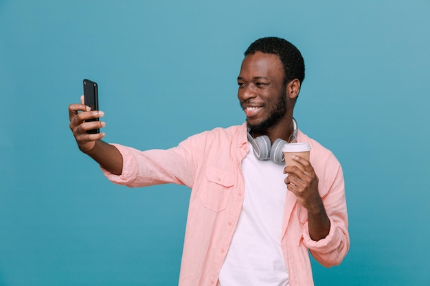 Sonriente sosteniendo una taza de café con teléfono joven afroamericano con auriculares en el cuello aislado sobre fondo azul.