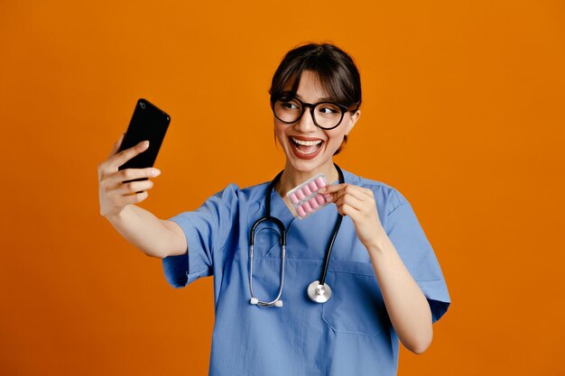 Sonriente, tómese un selfie sosteniendo píldoras, una joven doctora usando un estetoscopio uniforme aislado en un fondo naranja