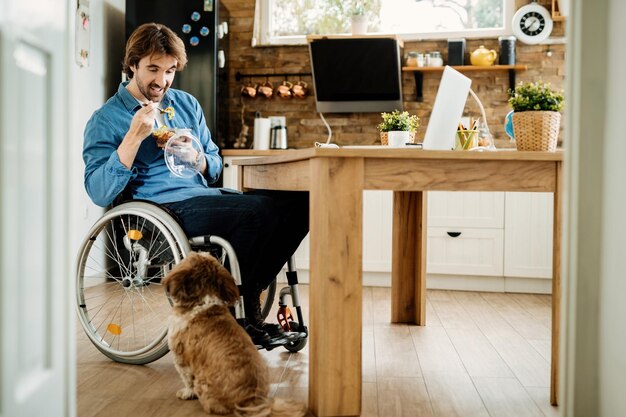 Sonriente trabajador independiente discapacitado disfrutando del almuerzo con su perro mientras trabaja en casa