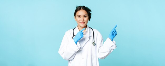 Sonriente trabajador sanitario asiático con guantes estériles y uniforme apuntando a la derecha demostrando el fondo azul del anuncio del hospital gráfico