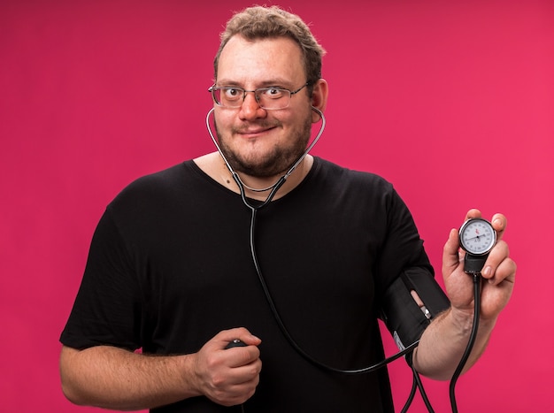 Sonriente varón enfermo de mediana edad midiendo su propia presión con esfigmomanómetro aislado en la pared rosa