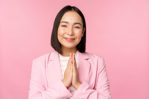 Sonriente vendedora asiática dama corporativa en traje agradeciendo tomarse de la mano en una bolsa gesto de gratitud de pie sobre fondo rosa