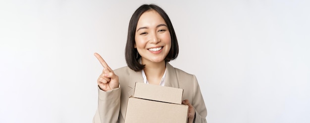 Sonriente vendedora asiática señalando con el dedo el espacio de la copia sosteniendo cajas con orden de entrega de pie sobre fondo blanco.