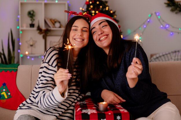 Sonrientes chicas guapas con gafas de reno y gorro de Papá Noel sosteniendo bengalas sentados en sillones y disfrutando de la Navidad en casa