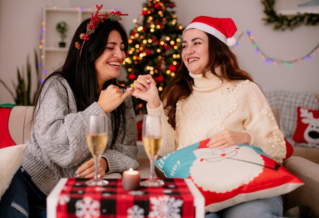 Sonrientes chicas guapas con gorro de Papá Noel cruzan sus deditos sentados en sillones y disfrutando de la Navidad en casa