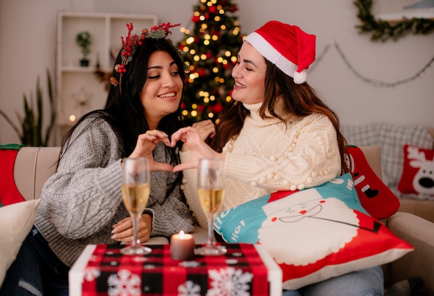 Sonrientes chicas guapas con sombrero de santa gesticulando signo de corazón juntos sentados en sillones y disfrutando de la Navidad en casa