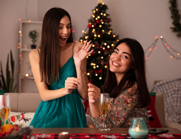 Sonrientes chicas guapas sostienen bengalas y miran a la cámara sentado en la mesa y disfrutando de la Navidad en casa
