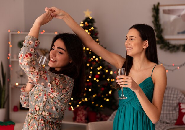 Sonrientes chicas guapas sostienen copas de champán y bailan juntos disfrutando de la Navidad en casa
