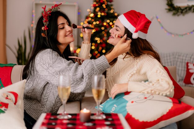 Sonrientes chicas guapas sostienen pinceles en polvo maquillándose mutuamente sentados en sillones y disfrutando de la Navidad en casa