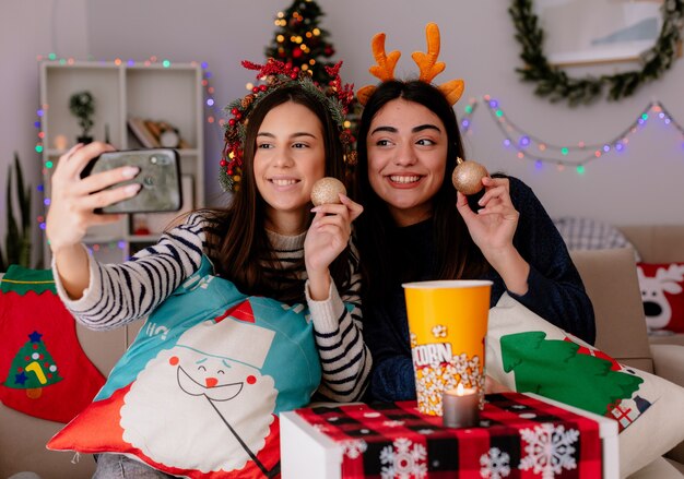 Sonrientes chicas jóvenes bonitas con corona de acebo y diadema de renos sostienen adornos de bolas de vidrio y toman selfie sentados en sillones y disfrutando de la Navidad en casa