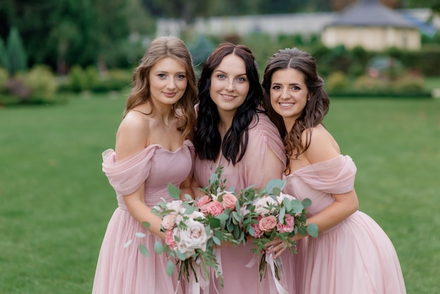 Sonrientes damas de honor atractivas vestidas con vestidos de color rosa pálido con tiernos ramos de rosas rosadas están sonriendo