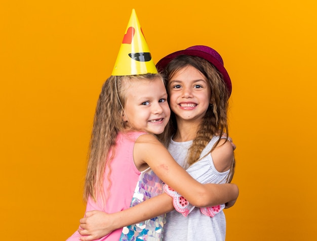 Sonrientes niñas bonitas con sombreros de fiesta abrazándose unos a otros aislados en la pared naranja con espacio de copia