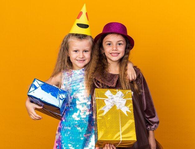 Sonrientes niñas bonitas con sombreros de fiesta sosteniendo sus cajas de regalo aisladas en la pared naranja con espacio de copia