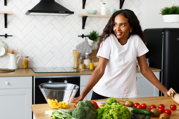 Sonrió hermosa mujer mulata está de pie cerca de la mesa llena de verduras frescas en la cocina moderna