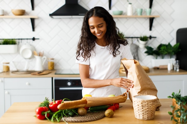 Sonrió hermosa mujer mulata está poniendo paquetes con comida sobre la mesa en la cocina moderna