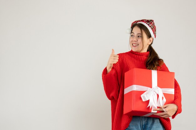 Sonrió linda chica con gorro de Papá Noel sosteniendo presente haciendo pulgar arriba firmar de pie en blanco