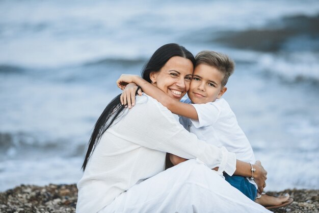 Sonrió madre e hijo abrazados y mirando directamente, sentados en la playa rocosa cerca del mar tormentoso