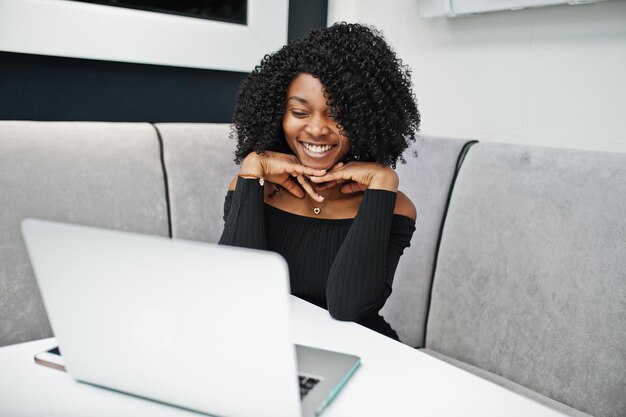 Sonrió a la moda joven y hermosa mujer de negocios afroamericana con peinado afro en elegante negro sentado y trabajando en la computadora portátil