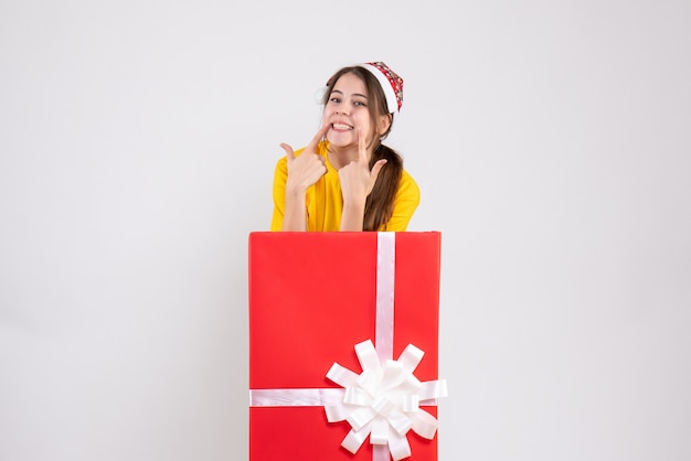 Sonrió niña con gorro de Papá Noel de pie detrás de un gran regalo de Navidad en blanco