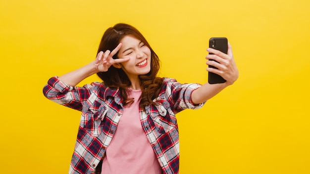 Sonrisa adorable mujer asiática haciendo selfie foto en smartphone con expresión positiva en ropa casual y mirando a cámara sobre pared amarilla. Feliz adorable mujer alegre disfruta el éxito.