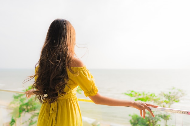 La sonrisa asiática joven hermosa de la mujer del retrato feliz y se relaja en el balcón al aire libre con la playa y el oce del mar