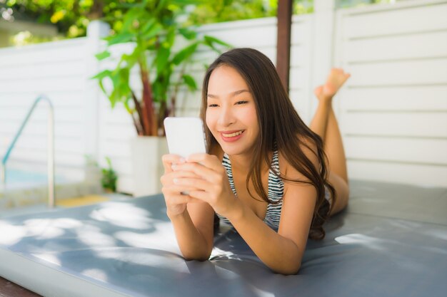 La sonrisa asiática joven hermosa de la mujer del retrato feliz se relaja con el teléfono móvil alrededor de piscina