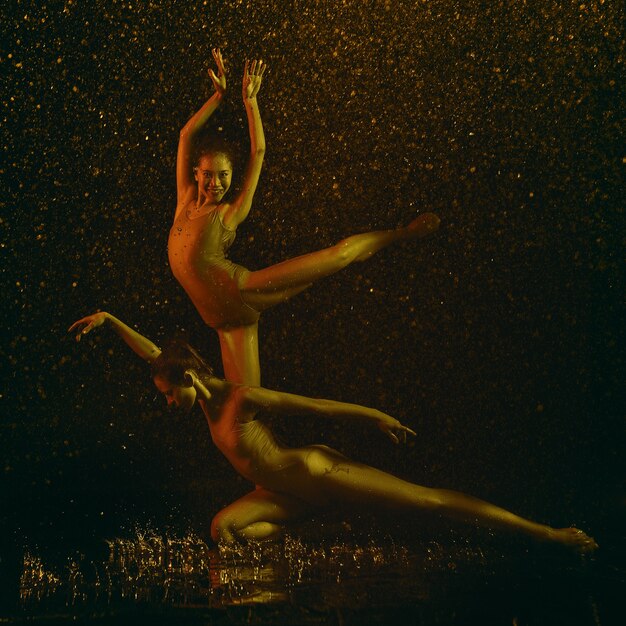 Sonrisa. Dos jóvenes bailarinas de ballet bajo gotas de agua y spray. Modelos caucásicos y asiáticos bailando juntos en luces de neón. Concepto de ballet y coreografía contemporánea. Foto de arte creativo.