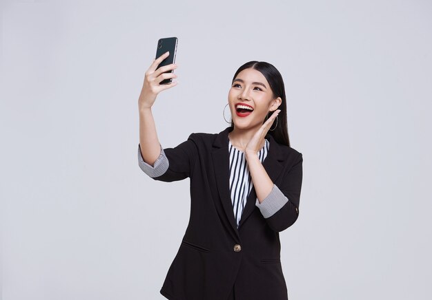 Sonrisa de empresaria asiática de cara feliz y amigable en traje formal que usa teléfono inteligente tiene una videollamada sobre fondo blanco foto de estudio.