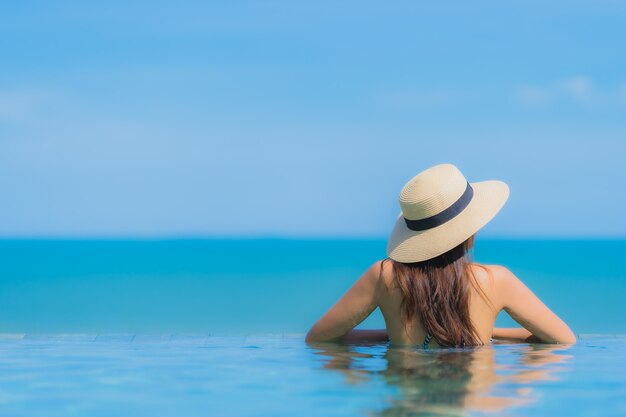 La sonrisa feliz de la mujer asiática joven hermosa del retrato se relaja en piscina en la playa neary del océano del mar del centro turístico del hotel en el cielo azul