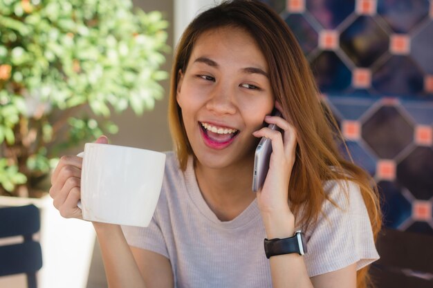 Sonrisa feliz las mujeres de negocios asiáticas que usan el teléfono celular que habla que se sienta en café y que sostiene una taza de coffe