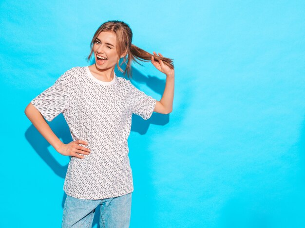 Sonrisa femenina positiva Modelo divertido que presenta cerca de la pared azul en estudio Jugando con el pelo y los guiños