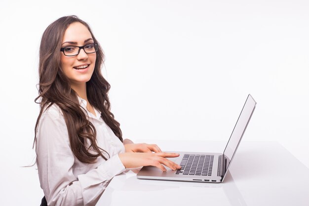 Sonrisa hermosa de la mujer de negocios que se sienta en el escritorio que trabaja usando la computadora portátil que mira la pantalla, escritura a mano, aislada sobre la pared blanca