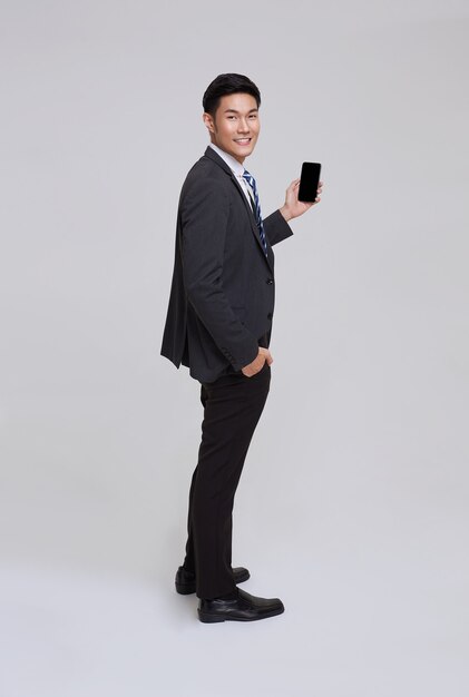 Sonrisa del hombre de negocios asiático de la cara hermosa y amistosa en traje formal que usa smartphone en tiro del estudio del fondo blanco