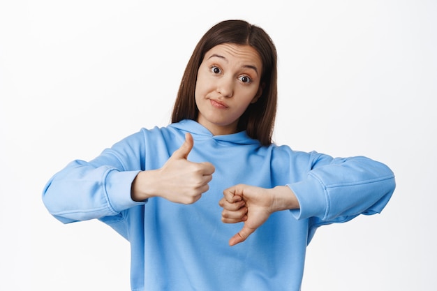 Sonrisa de mujer joven, muestra disgusto, gesto de pulgar hacia arriba y pulgar hacia abajo, calificación media, resultado promedio, de pie en sudadera con capucha contra la pared blanca