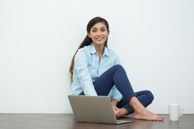 Sonrisa de la mujer joven con ordenador portátil y té en el piso