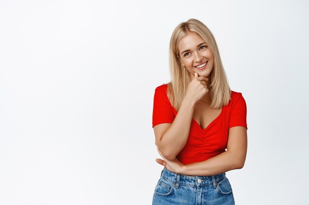 Las sonrisas de una chica rubia glamurosa se ven coquetas ante los soportes de la cámara en top rojo recortado y jeans contra fondo blanco