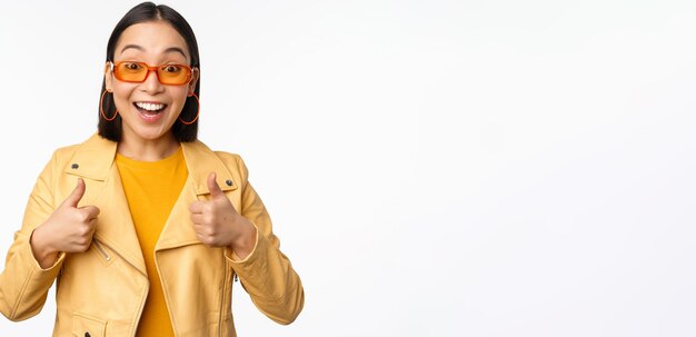 Las sonrisas entusiastas de las niñas asiáticas muestran los pulgares hacia arriba y se ven felices diciendo que sí, aprueban y recomiendan algo de pie sobre un fondo blanco