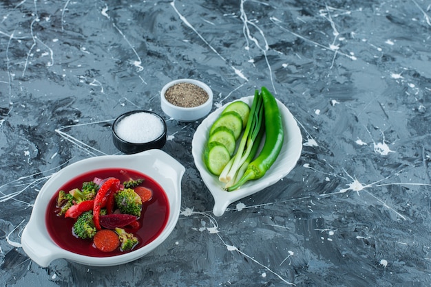 Sopa de borsch en un cuenco junto a verduras y especias, sobre fondo azul.