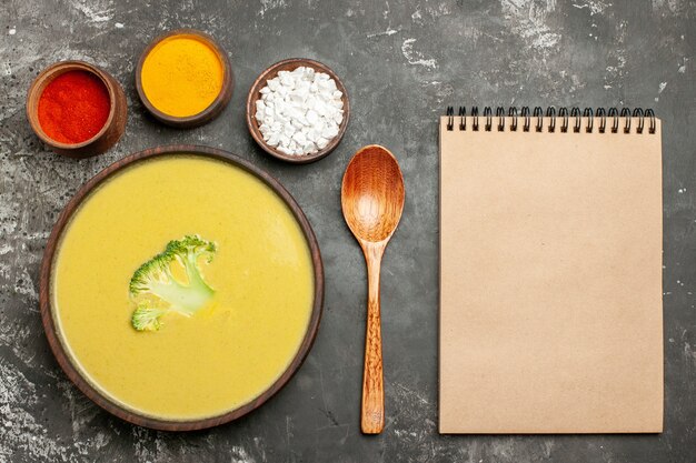 Sopa cremosa de brócoli en un cuenco marrón, cuchara de diferentes especias y cuaderno sobre mesa gris