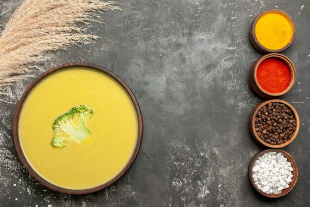 Sopa cremosa de brócoli en un recipiente marrón y diferentes especias en mesa gris