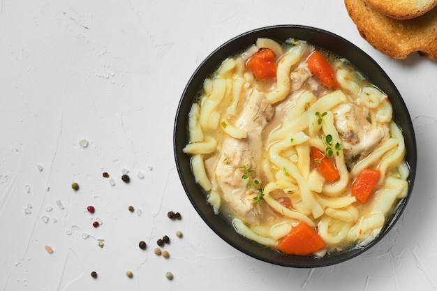 Sopa de fideos de pollo casera con verduras y especias en un recipiente, vista superior, fondo de hormigón blanco.